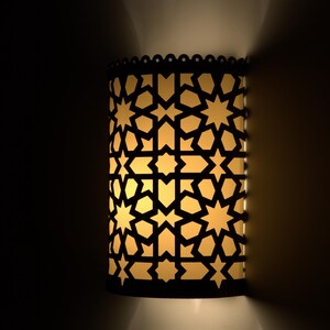 May include: A cylindrical wall sconce featuring a dark, laser-cut metal overlay with a repeating geometric star and polygon pattern. The fixture emits a warm, soft yellow light through the cutouts, casting a glow against the wall surface.