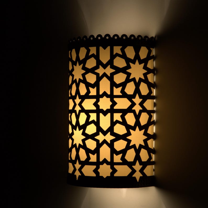 May include: A cylindrical wall sconce featuring a dark, laser-cut metal overlay with a repeating geometric star and polygon pattern. The fixture emits a warm, soft yellow light through the cutouts, casting a glow against the wall surface.