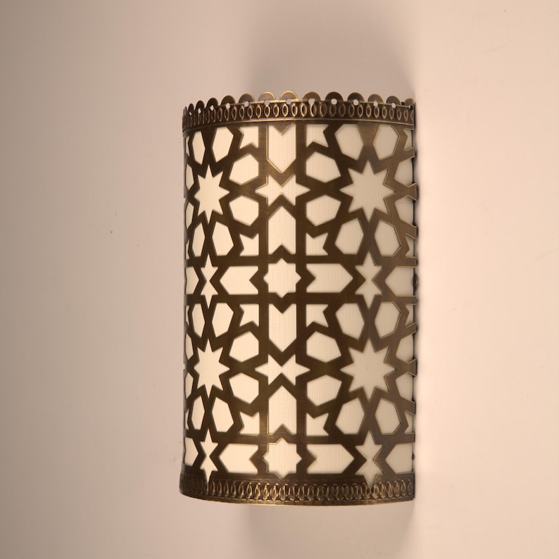 May include: A semi-cylindrical wall sconce featuring a metallic bronze-toned frame with an intricate, laser-cut geometric star and polygon pattern. The frame overlays a white, translucent panel that diffuses light, with scalloped edges.
