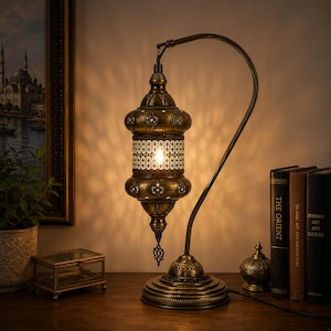 May include: A brass-toned metal table lamp with an arched stand and a hanging lantern-style shade. The shade features intricate cut-out patterns that cast light and shadows, with a central cylindrical section and decorative metal finials.