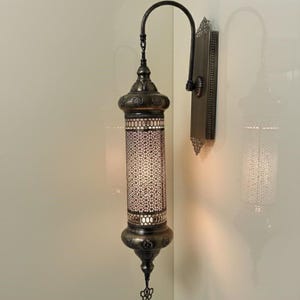Puede incluir: Un aplique de pared con una pantalla cil&iacute;ndrica de metal perforado con un intrincado patr&oacute;n geom&eacute;trico. La l&aacute;mpara tiene un acabado en lat&oacute;n antiguo oscuro, un brazo curvo y una placa rectangular con bordes de filigrana decorativa.