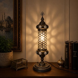 May include: A cylindrical table lamp with an antique bronze finish and a honeycomb-patterned glass shade. The lamp features a decorative metal top finial and a circular base, emitting a warm glow that casts intricate shadows on the wall.