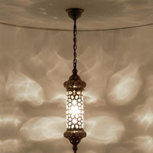 May include: A bronze pendant light with a cylindrical shade. The shade has a star-shaped pattern, allowing light to filter through. The fixture hangs from a chain and is suspended from a ceiling mount.