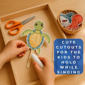 May include: A wooden tray contains a green turtle cutout on a stick, orange scissors, and a small basket with shark and octopus cutouts. Text on a blue square reads "Cute cutouts for the kids to hold while singing."