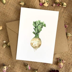 May include: A watercolour greeting card showing a celeriac root vegetable with green leaves. The card is on a white background, with a brown envelope behind it. The image is set against a hessian background.