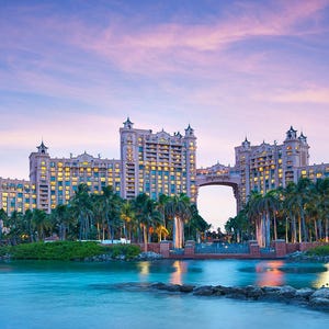 May include: A scenic view of a large resort complex at dusk. The buildings are a light pink color with many windows, connected by an archway. Palm trees line the waterfront, and the sky is a mix of pink and blue.
