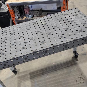 Puede incluir: Una mesa de trabajo metálica rectangular grande con una superficie perforada y un marco gris oscuro. La mesa tiene cuatro patas con ruedas para la movilidad. Las patas son de color gris claro con detalles en negro. Diseñada para uso industrial.