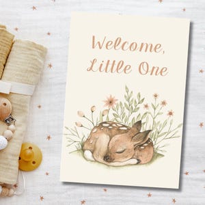 May include: A greeting card with the text "Welcome, Little One" in a soft brown font. The card features a watercolor illustration of a sleeping fawn surrounded by greenery and flowers. The card has a cream background.