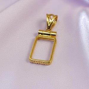May include: A gold-colored pendant with a rectangular frame and a decorative bail. The pendant is set against a soft, light purple fabric background. The frame has a textured edge.