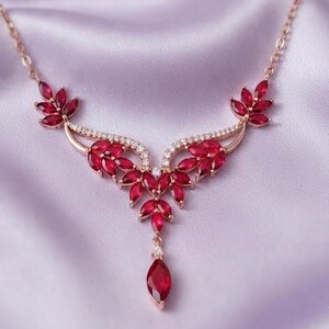 Könnte beinhalten: Eine Halskette aus Roségold mit einem zentralen Anhänger aus tiefroten Steinen im Marquise-Schliff und kleinen klaren Akzentsteinen. Das Design zeigt ein symmetrisches blattartiges Muster mit einem einzelnen tropfenförmigen roten Stein.