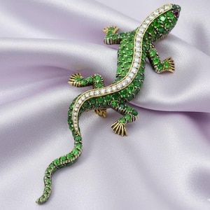 May include: A jeweled lizard brooch with a green body, a white diamond-lined spine, and gold-colored feet. The lizard's body is covered in green gemstones, and a small red gemstone is set at the head.