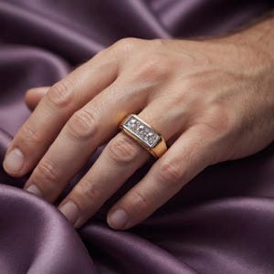 May include: A gold ring featuring a rectangular setting with three clear, faceted stones arranged in a horizontal row. The band is smooth and polished, with a wide, flat profile that sits securely on the finger, showcasing a metallic finish.