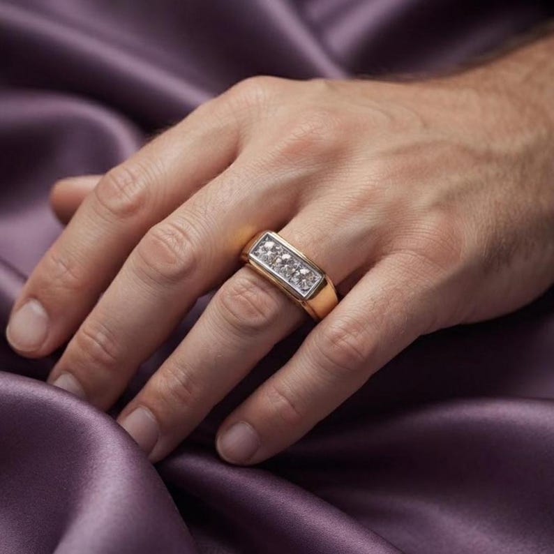 May include: A gold ring featuring a rectangular setting with three clear, faceted stones arranged in a horizontal row. The band is smooth and polished, with a wide, flat profile that sits securely on the finger, showcasing a metallic finish.