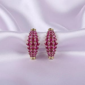 May include: A pair of gold-toned hoop earrings, each encrusted with numerous oval-shaped, deep pink gemstones. The earrings are set against a soft, lavender-colored satin background, creating a luxurious and elegant aesthetic.