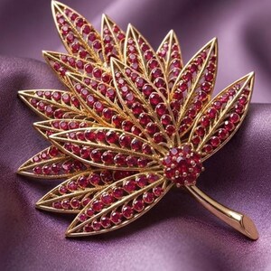 May include: A gold-toned brooch shaped like a multi-layered leaf, featuring a detailed openwork metal frame densely set with numerous small, round, deep red stones. The design includes a central cluster of red stones at the base of the stem.