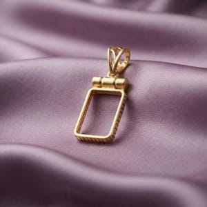 Puede incluir: Un colgante rectangular de tono dorado con un centro hueco y un borde exterior texturizado. El colgante incluye una reasa decorativa calada en la parte superior, diseñada para sujetarse a una cadena o cordón, con un acabado metálico pulido.