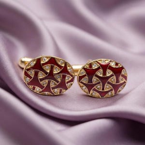 Könnte beinhalten: Ein Paar ovale Manschettenknöpfe aus Gold mit einer tiefroten Emaille-Einlage in einem sich wiederholenden geometrischen Muster, akzentuiert durch kleine klare Steine, die in das Design eingebettet sind. Polierte goldfarbene Metalloberfläche.