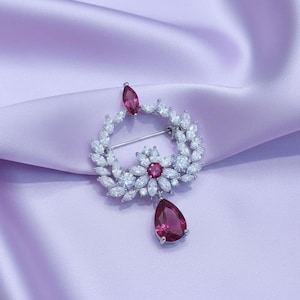 May include: A silver-toned brooch with a floral and leaf design, featuring clear and ruby-colored gemstones. The brooch has a central flower design with a ruby center and a teardrop-shaped ruby pendant. The brooch is set against a lavender satin background.
