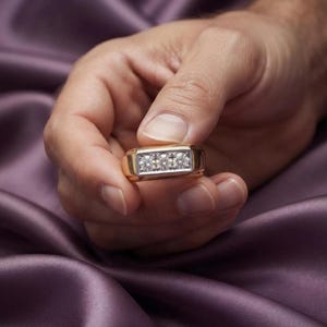 May include: A gold ring featuring a rectangular silver-toned setting that holds three round, clear, faceted stones arranged in a horizontal row. The band is smooth and polished, showcasing a classic design with a metallic finish and clear gems.
