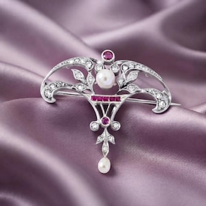 Op de afbeelding: Een zilverkleurige broche met een centrale witte parel, geaccentueerd met kleine heldere stenen en rechthoekige rode stenen. Het metaalwerk toont een ingewikkeld, symmetrisch, bladachtig ontwerp met een bungelende parel aan de onderkant.