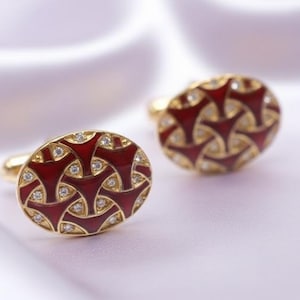 Puede incluir: Un par de gemelos dorados con un diseño de esmalte rojo y pequeñas piedras preciosas transparentes. Los gemelos ovalados tienen un patrón geométrico. El fondo es de tela blanca suave.