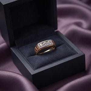 May include: A gold ring featuring a rectangular setting with three round, clear, faceted stones arranged in a horizontal row. The ring is displayed inside a dark, square-shaped jewelry box with a soft, dark-colored interior lining.