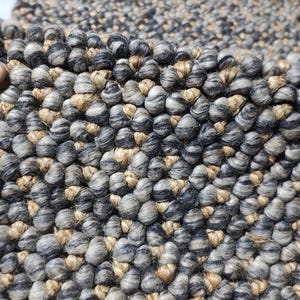 May include: A close-up shot of a textured rug, featuring a pattern of grey, black, and tan woven fibres. The rug's surface is composed of tightly packed, rounded elements, giving it a tactile feel. The colour scheme is neutral, with a blend of light and dark shades.