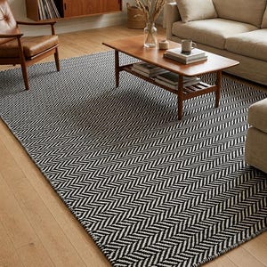 May include: A large rectangular area rug featuring a repeating black and white herringbone pattern. The rug is laid flat on a light-colored wooden floor, covering a significant portion of the space beneath a wooden coffee table and surrounding furniture.