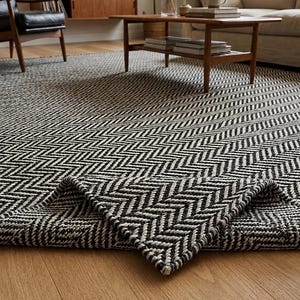 May include: A large area rug featuring a high-contrast black and white herringbone pattern woven with a thick, textured corded material. The rug is laid flat on a wooden floor with one corner folded over to reveal the underside and thickness.