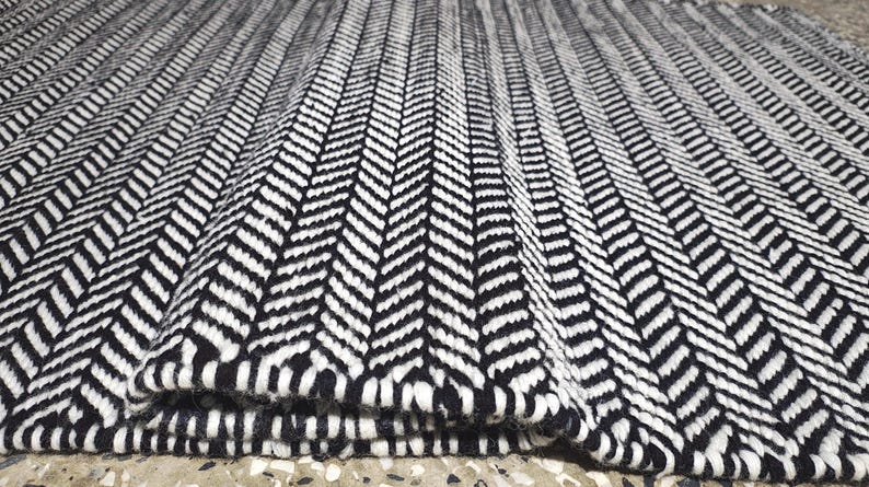 May include: A rectangular area rug featuring a high-contrast black and white herringbone pattern. The woven textile displays a repeating V-shaped chevron design across the entire surface, with one corner folded over to reveal the thickness of the rug.