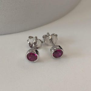 May include: A pair of stud earrings with a silver finish. Each earring has a small, oval-shaped, deep pink gemstone set within a bezel. The earrings are displayed on a white surface.