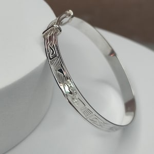 May include: A silver bangle bracelet with engraved geometric and floral patterns. The bracelet has a clasp closure and is displayed on a white surface. The design is detailed and the metal has a polished finish.