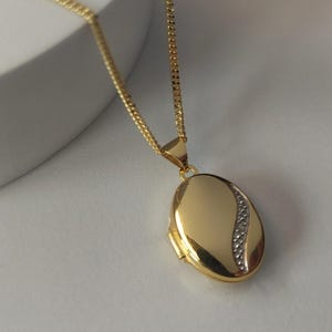 May include: A gold locket necklace with a fine chain. The oval pendant has a curved design with a line of small, sparkling stones. The necklace is displayed on a white surface.