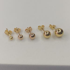 May include: Five pairs of gold ball stud earrings in varying sizes are displayed on a white surface. The earrings have a classic design, with a small post and a round ball. The earrings are made of a shiny gold material.
