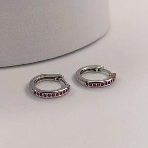 May include: Two silver hoop earrings, each featuring a line of small, red gemstones. The earrings are presented on a white surface, highlighting their shiny metal and the vivid colour of the stones.