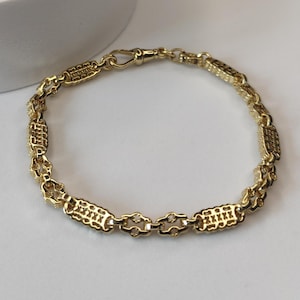 May include: A gold bracelet with a decorative chain design. The bracelet features rectangular and oval links connected by smaller, decorative links. The clasp is a simple hook and eye closure. The jewellery is displayed on a white surface.