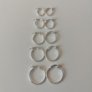 May include: Six pairs of small, silver hoop earrings are displayed in rows on a light grey surface. The earrings are of varying sizes, featuring a simple, classic design. The metal has a shiny, reflective quality.