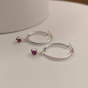 May include: A pair of silver hoop earrings, each with a small, dangling red gemstone. The earrings are presented on a white surface, against a soft, neutral background. The gemstones are a deep red colour.