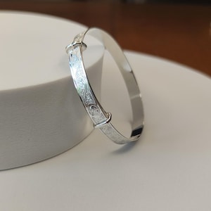 May include: A silver adjustable bangle. The bracelet has an engraved pattern on the adjustable section. It is displayed on a white surface. The bangle is made of a shiny, reflective metal.