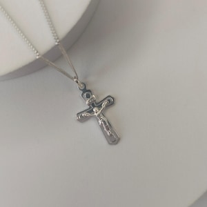 May include: A silver crucifix pendant with a detailed depiction of Jesus Christ on the cross. The pendant hangs from a delicate silver chain. The cross has a dark border and a detailed figure of Jesus.