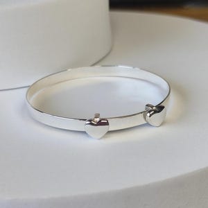 May include: A polished silver bangle bracelet featuring two small, smooth heart-shaped charms attached to the band. The bracelet has a clean, reflective metallic finish and a simple, circular design that maintains a consistent width throughout.