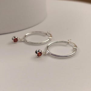 May include: Two silver hoop earrings, each with a small, round, red gemstone pendant. The earrings are shown against a white backdrop. The hoops are circular and have a simple design.