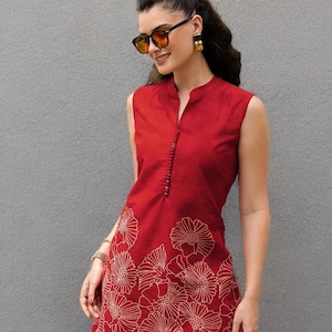 Könnte beinhalten: Ärmellose rote Tunika mit Stehkragen und floralem Design. Die Tunika hat eine Knopfleiste und ein Blumenmuster in einem helleren Rotton. Die Person trägt Sonnenbrille und Ohrringe.
