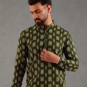 Puede incluir: Camisa de manga larga verde oliva con cuello mandarín y cierre de botones. La camisa presenta un patrón geométrico repetitivo en tonos beige y marrón. El modelo lleva la camisa con pantalones beige.