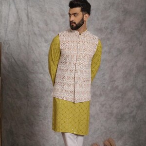 May include: A three-piece ensemble featuring a yellow kurta, white pants, and a cream-colored vest with a geometric pattern. The vest has a mandarin collar and button closure. The outfit is completed with brown sandals.