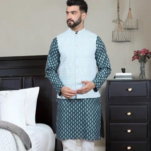 May include: A three-piece ensemble featuring a light blue vest over a patterned teal and black tunic and sleeves. The outfit is completed with white pants. The man is wearing brown sandals.
