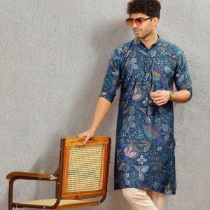 May include: A teal floral print kurta with cream-colored pants. The kurta features a button-down front and a mandarin collar. The outfit is paired with brown leather sandals. A wooden chair with a woven back is also in the image.