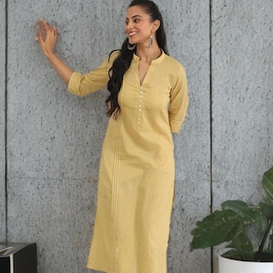 Può includere: Una donna indossa un kurta giallo a righe con pantaloni abbinati. Il kurta ha un colletto alla coreana e dettagli con bottoni. L'outfit è abbinato a tacchi beige. Una pianta in vaso è sullo sfondo.