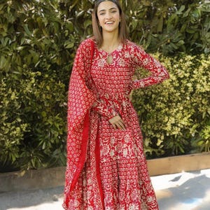 May include: A red long-sleeved dress with a matching scarf, featuring a white floral pattern. The dress has a keyhole neckline and a flowing skirt. The outfit is worn with clear sandals.