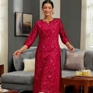 以下が含まれることがあります： マゼンタ色のチュニックに花の刺繍、クリーム色のパンツを組み合わせたスタイル。チュニックはVネックで、七分袖。装いを完成させるのは装飾されたフラットシューズ。背景にはグレーのソファと木製家具があります。